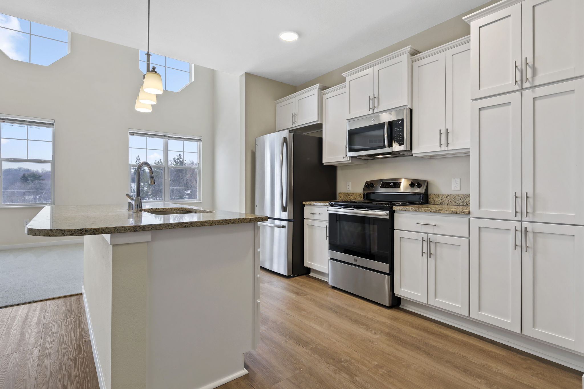 A kitchen setting with an island-counter near the middle of the room, with a sink in it. There are cabinets, an oven and stove, fridge, and microwave on the back-left wall.