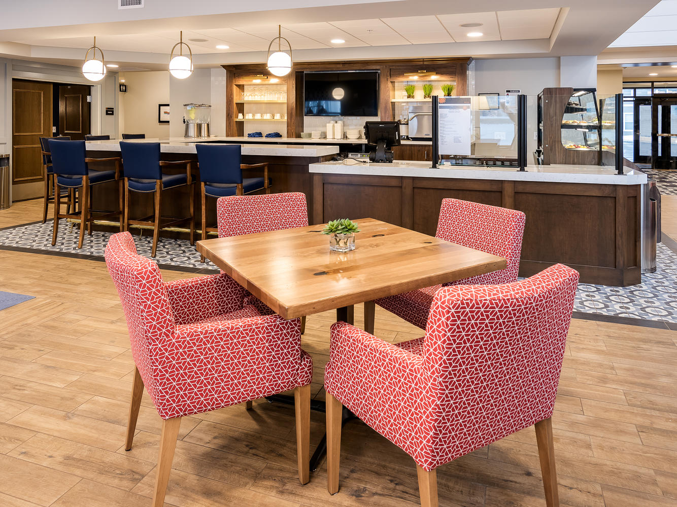 A wooden table surrounded by red and white chairs. There are blue chairs in front of a countertop in the back of the room.