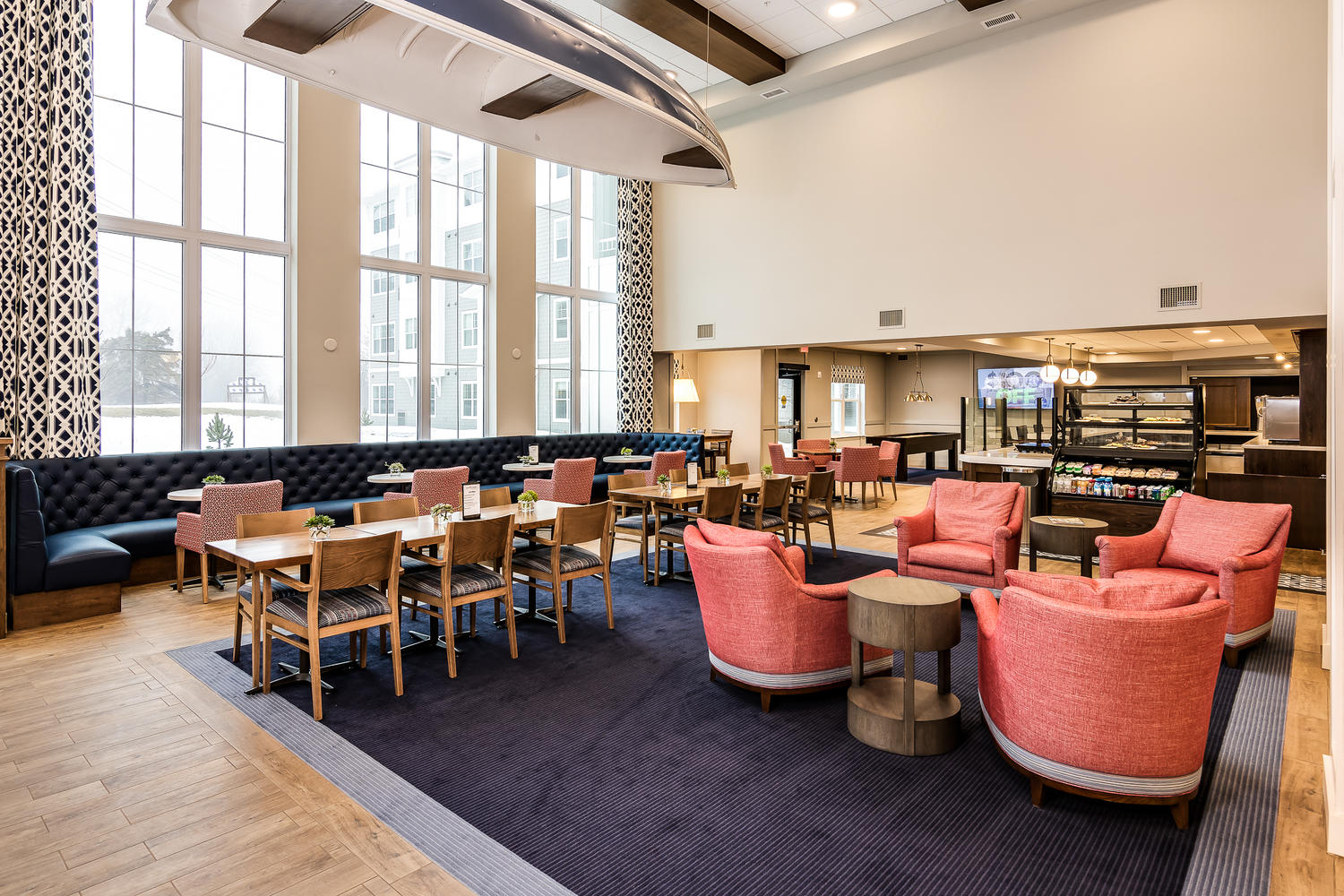 A dining room with red lounger chairs, and brown wooden chairs sitting around differently-sized tables. They are on blue carpet, in front of large windows on the back-wall.