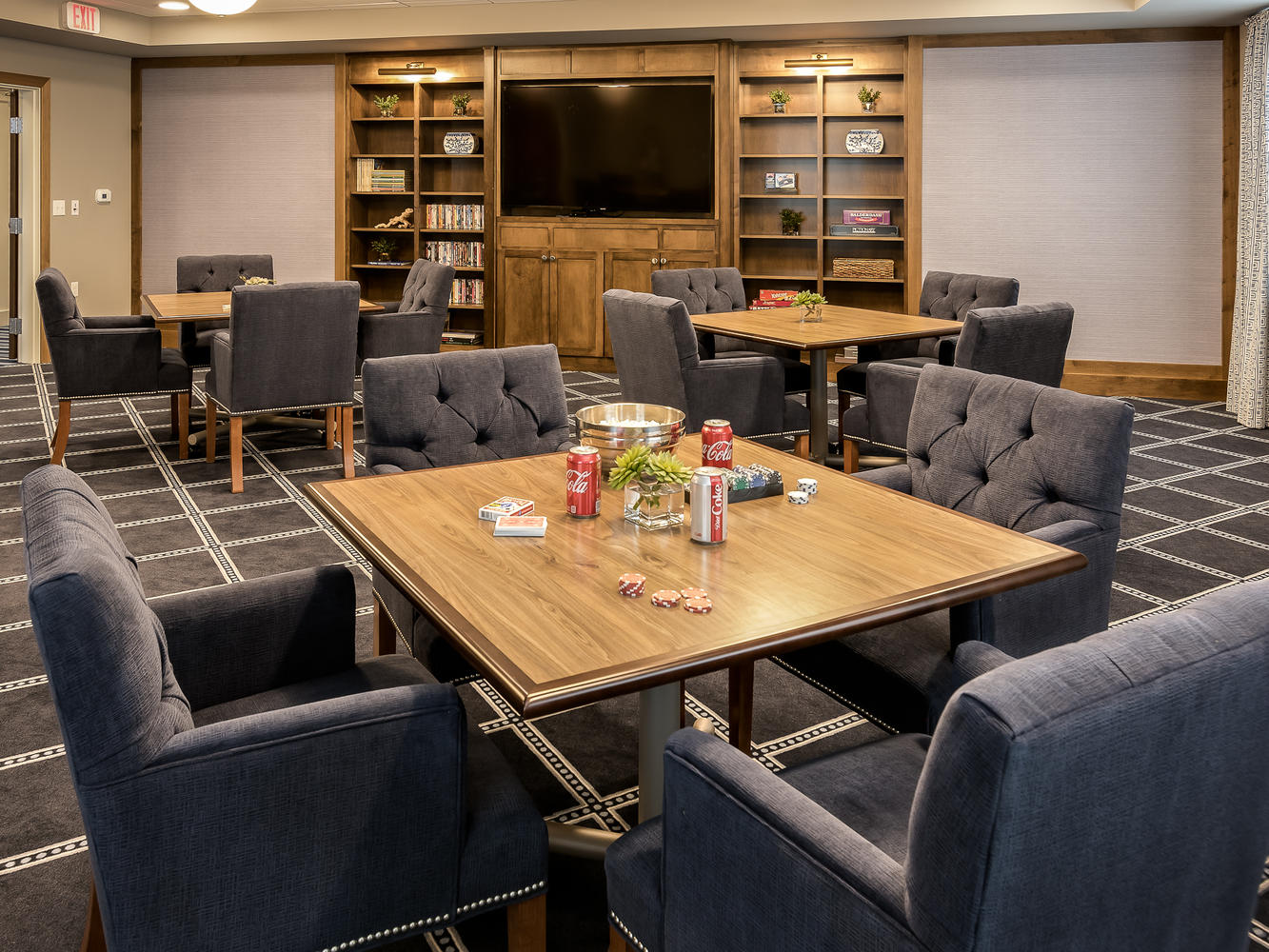 A few tables with soda, cards and poker chips on it. Surrounded by four gray chairs, and a tv and bookshelves near the back of the room.