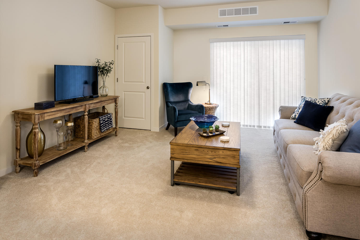 A living room with a tan couch, wooden table, blue chair in the corner, and tv sitting on a wooden table near the back wall.