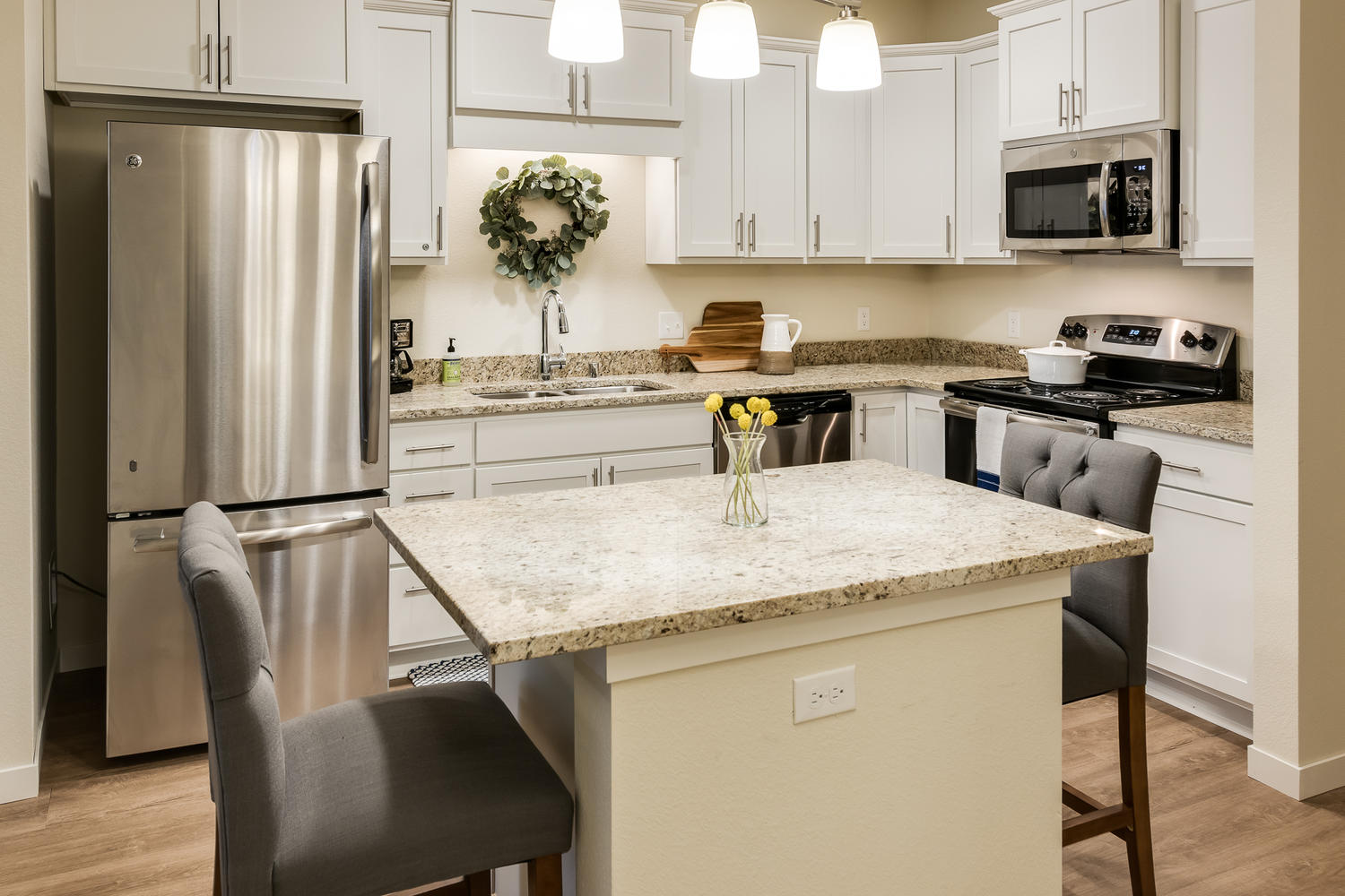 A table with two chairs on different sides of it, in front of a kitchen setting. There's a fridge, countertops, a sink, and cabinets behind the table.