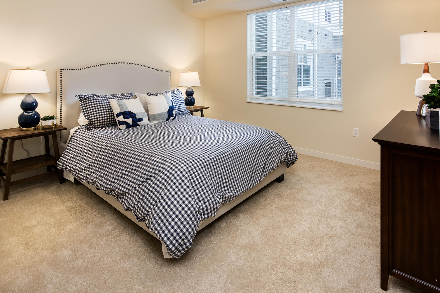 A large bed with a blue and white comforter sitting on the carpet floor. There are pillows on the bed, and a nightstand with a lamp on each side of the bed.