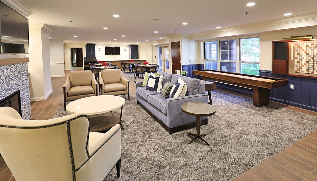 A living-room area with a gray sofa centered in the middle of the room, with a white table sitting in front of it, and chairs to the left and right. There is a TV mounted on the wall in front of the sofa.