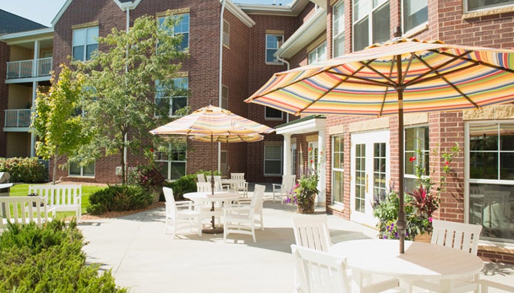 Multiple white tables sitting outside the entrance of a building, with four white chairs around each one. In the center of each table is a colorful umbrella blocking out some of the sun.