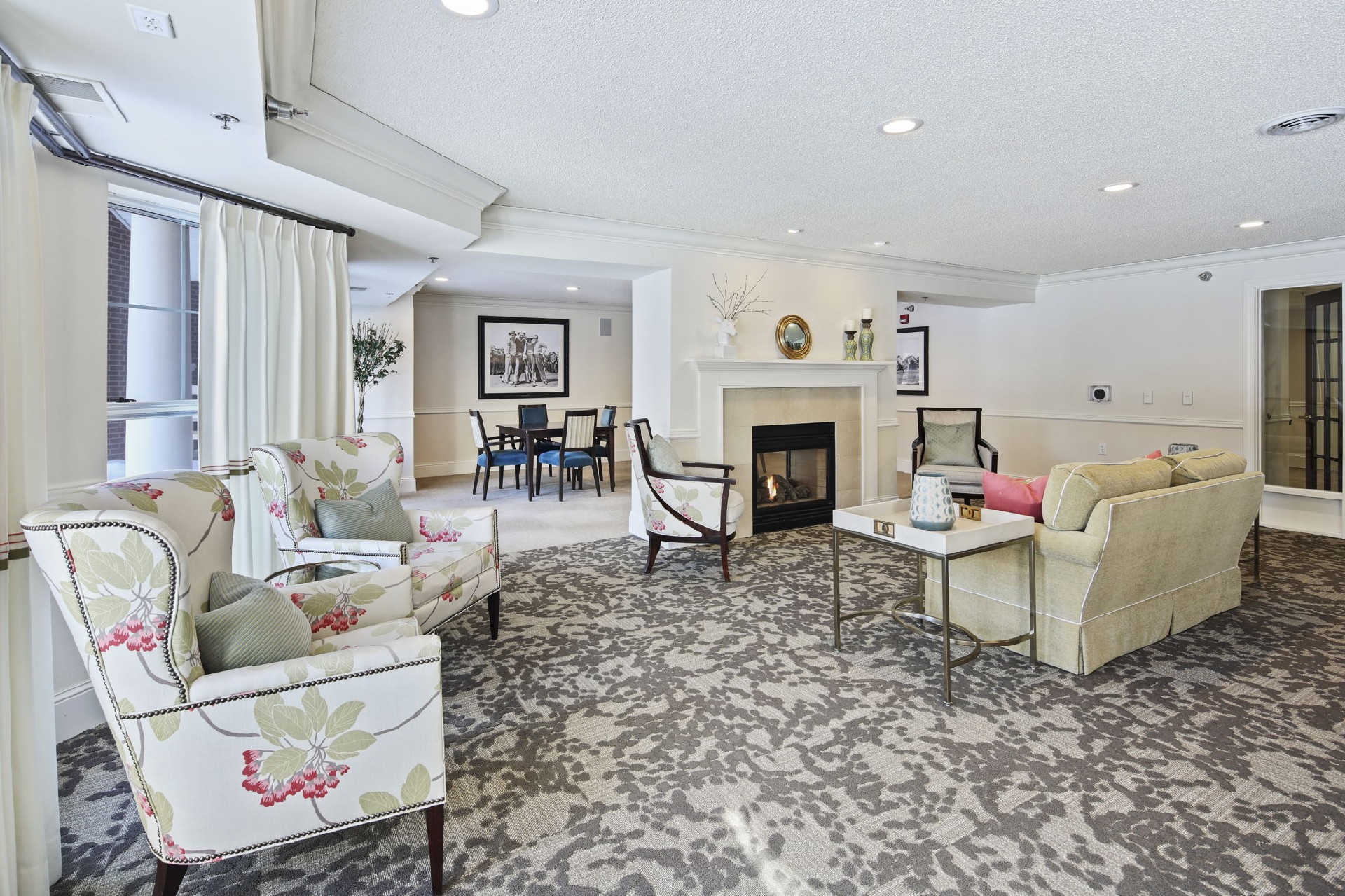 White room with a few chairs sitting against the wall, a soft sitting in the middle with a fireplace in front of it, and a dining room table with chairs near the back.