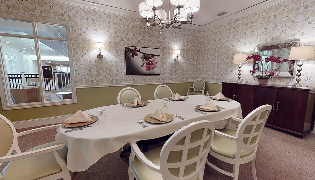 A long, white dining table with many chairs sitting around it. The table is set with plates, napkins, and silverware. There is a counter with a couple lamps on it near the back of the room.