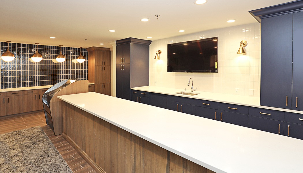 A long white countertop in front of a kitchen-like area with countertops, a sink, and a tv.