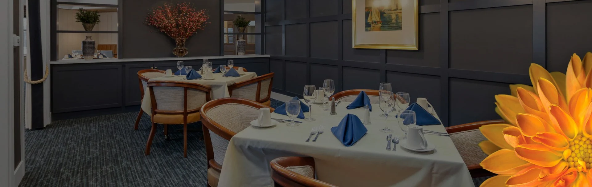 A cozy room with navy blue wallpaper & carpet. There are two tables each with four chairs around them, and they're set with glasses and silverware.