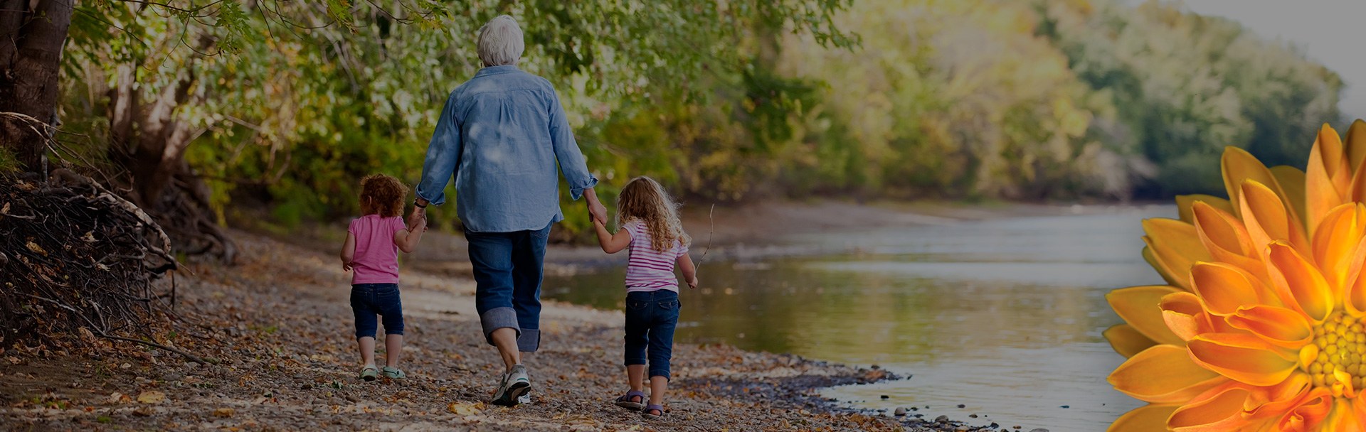 An older women holding the hands of two young girls, walking next to a small body of water to the right, and a forest to the left.