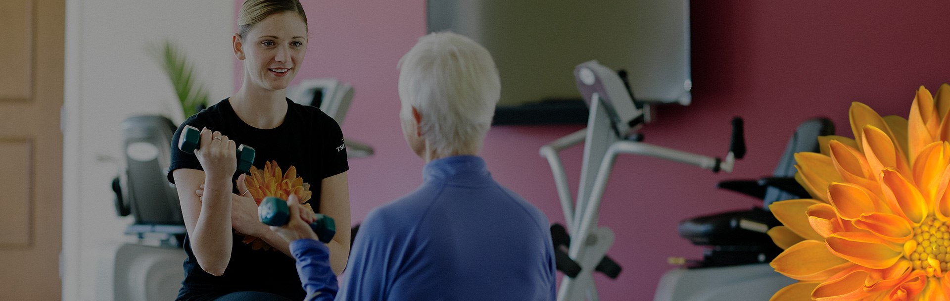 Girl lifting a weight in her right hand, sitting in front of an older women also lifting a weight. Both are in a fitness room with equipment sitting around the room.