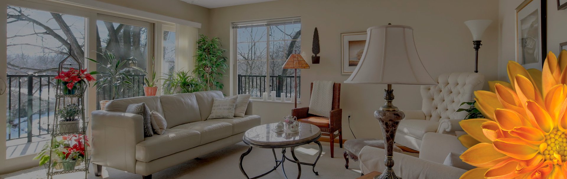 A living room setting with a tan couch, marble table, many lamps, flowers, and windows showing a snowy landscape.