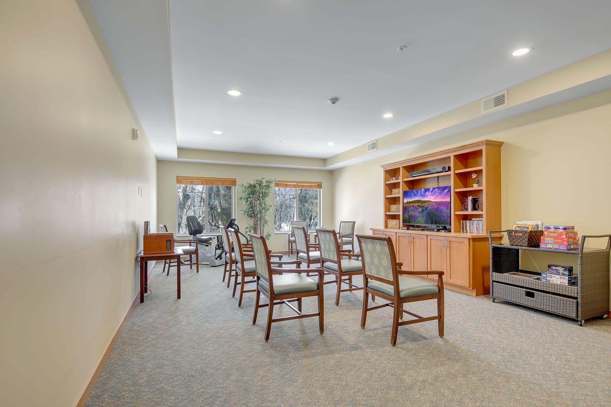 A room with gray chairs all facing towards a tv on a bookshelf up against the wall. There are board games on a cart next to the bookshelf, and windows on the back-left wall.