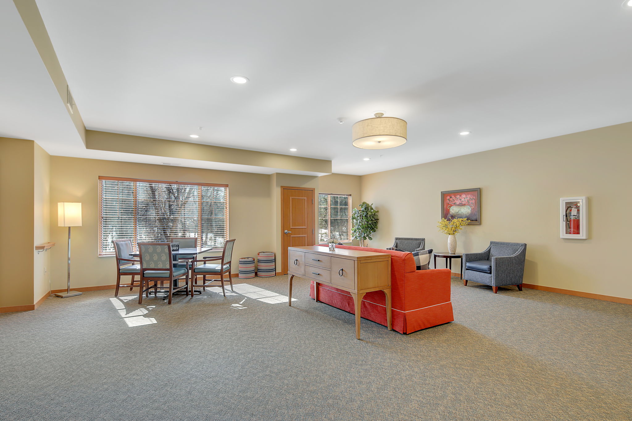 An open room with a red couch, a couple blue and white chairs, a table with chairs around it, a lamp in the corner, and a door near the back-right of the room. There are windows next to the door.