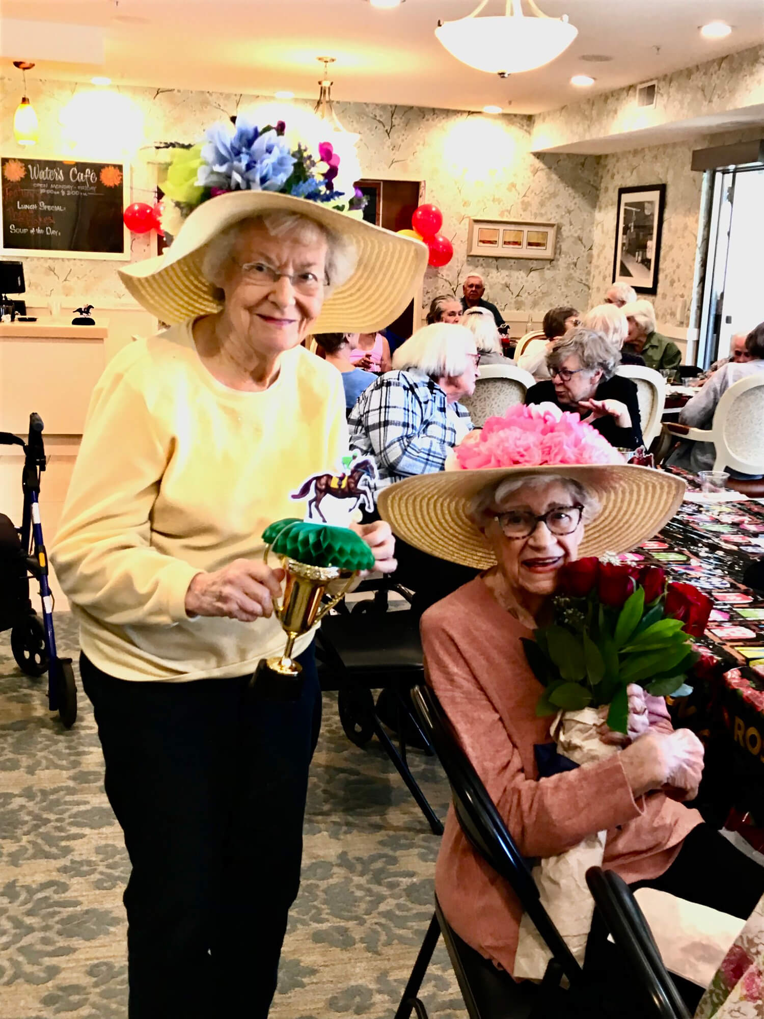 Two senior ladies wearing decorative hats with flowers and ribbons celebrate Derby Day, one holding a trophy and the other a bouquet of roses.