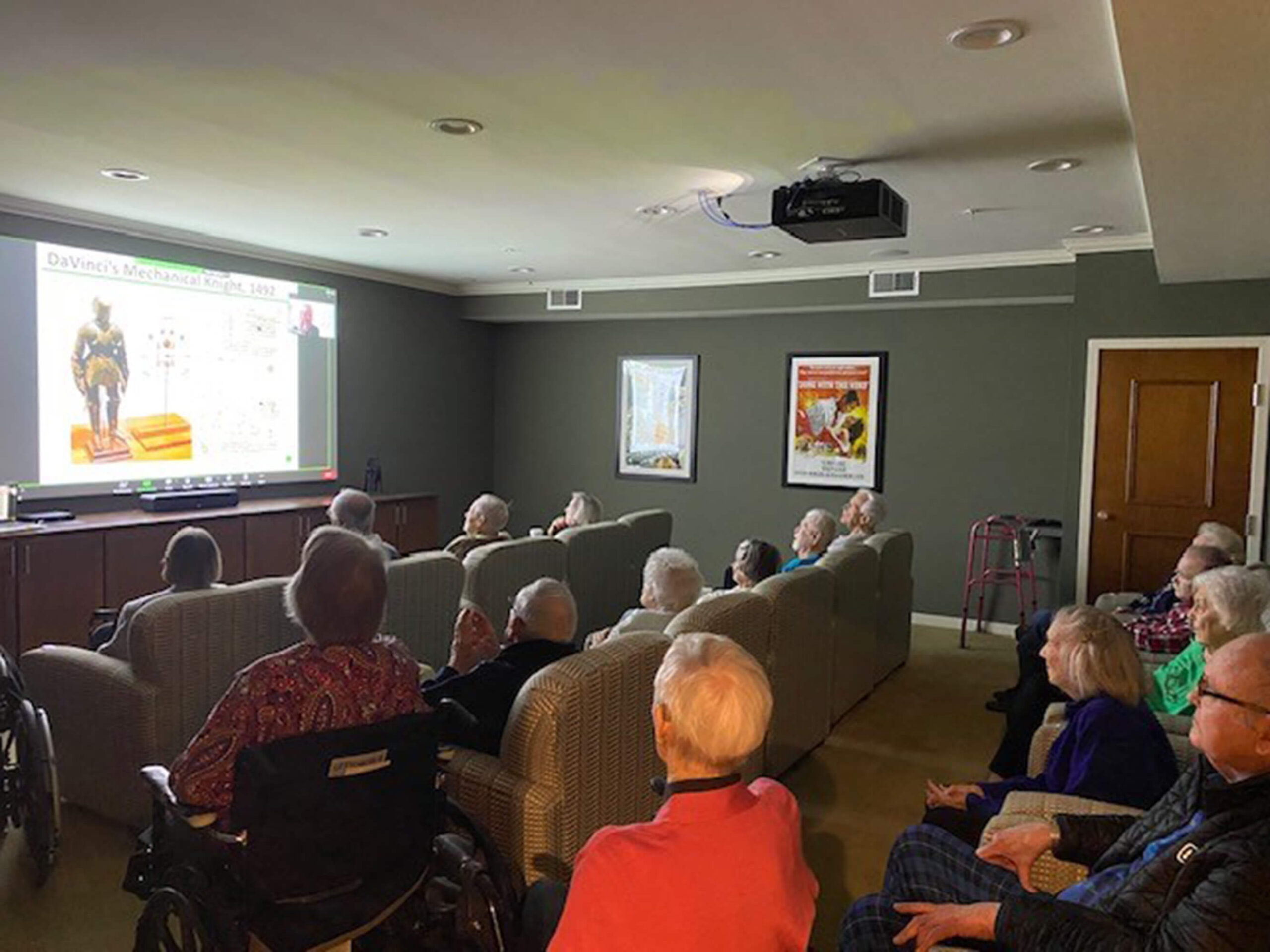 Group of seniors attentively watching an educational seminar in a darkened room, featuring a presentation on Da Vinci's mechanical knight.