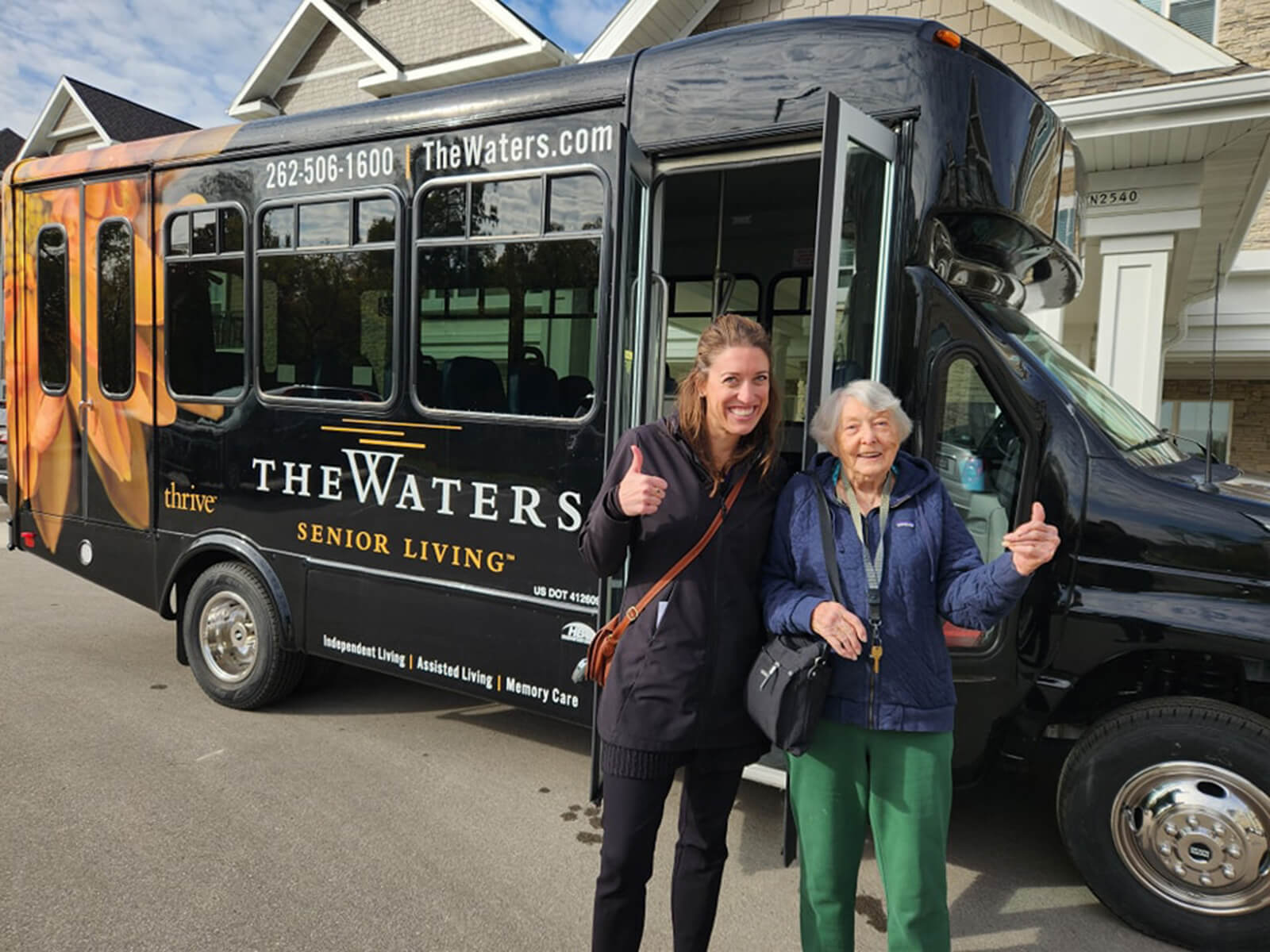 A joyful senior resident ready for an outing, standing beside The Waters of Pewaukee community bus, symbolizing active senior living.