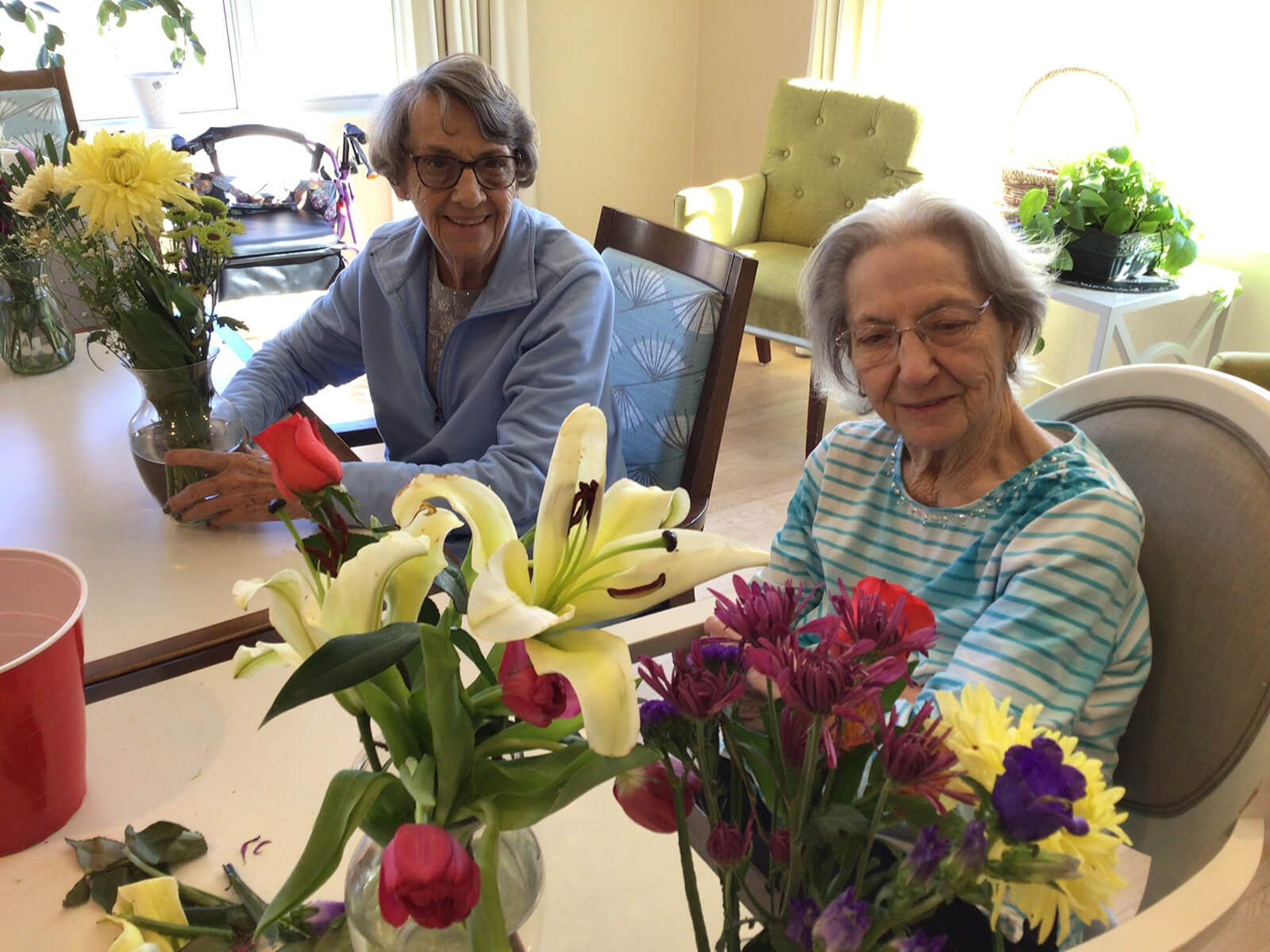 Two senior residents at The Waters on Mayowood smiling as they engage in a flower arrangement activity, enhancing community living.