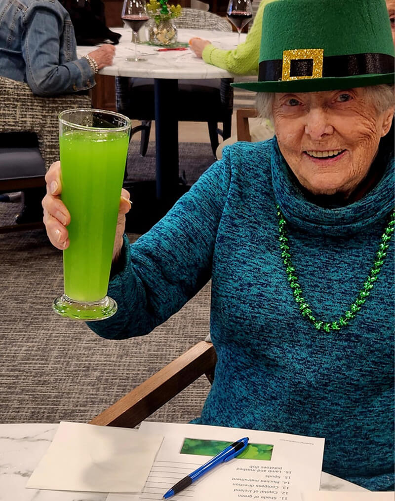 A senior resident at The Waters of Pewaukee celebrating St. Patrick's Day with a festive green hat and a toast with a vibrant green drink.